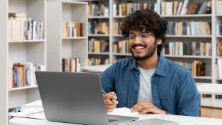 Smiling male student sitting in front of an open laptop in a library.