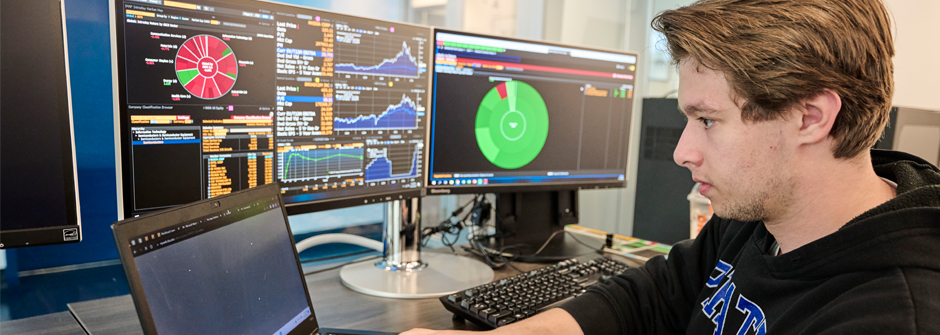 Image of a student sitting in front of two monitors with charts and pie graphs on the screen, with the student wearing a Seton Hall hoodie. 