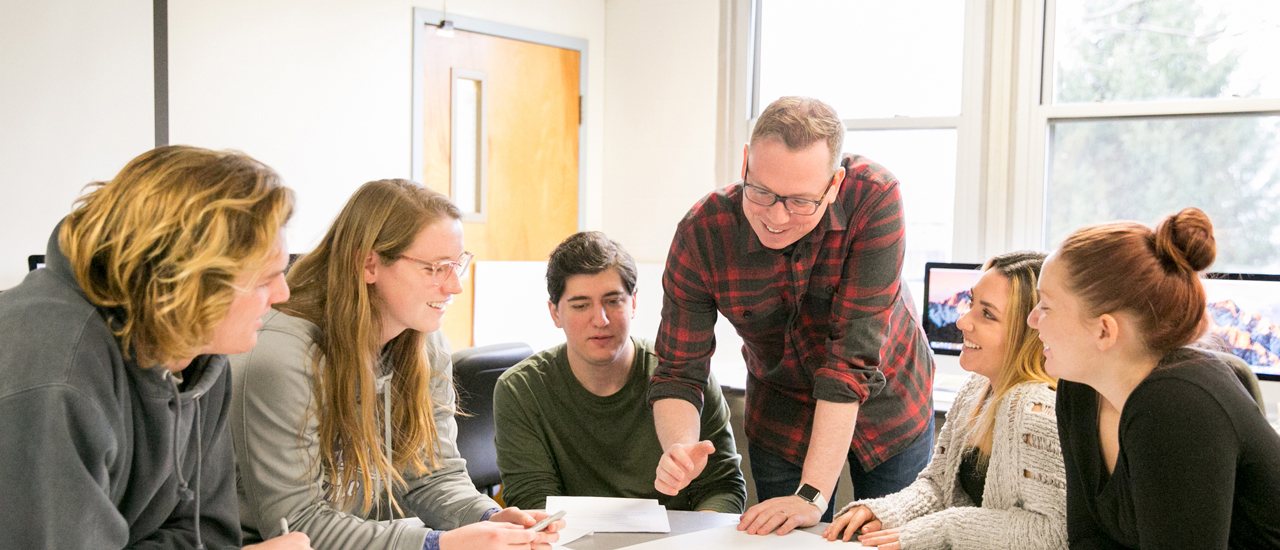 Image of guest Brian Meszaros with Seton Hall students collaborating on a project in front of a row of PC computers. 