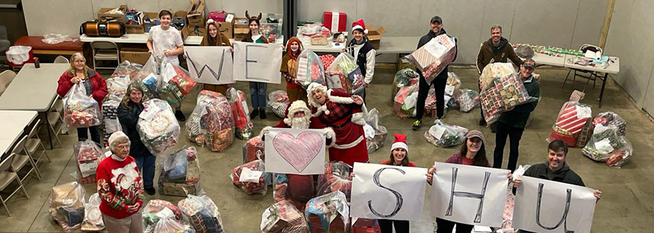 Toy recipients in a warehouse holding up a sign saying "I Love Seton Hall" and wearing santa hats. There are wrapped gifts at their feet for children. 