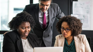 Women working on a computer with their manager looking over their shoulder. 